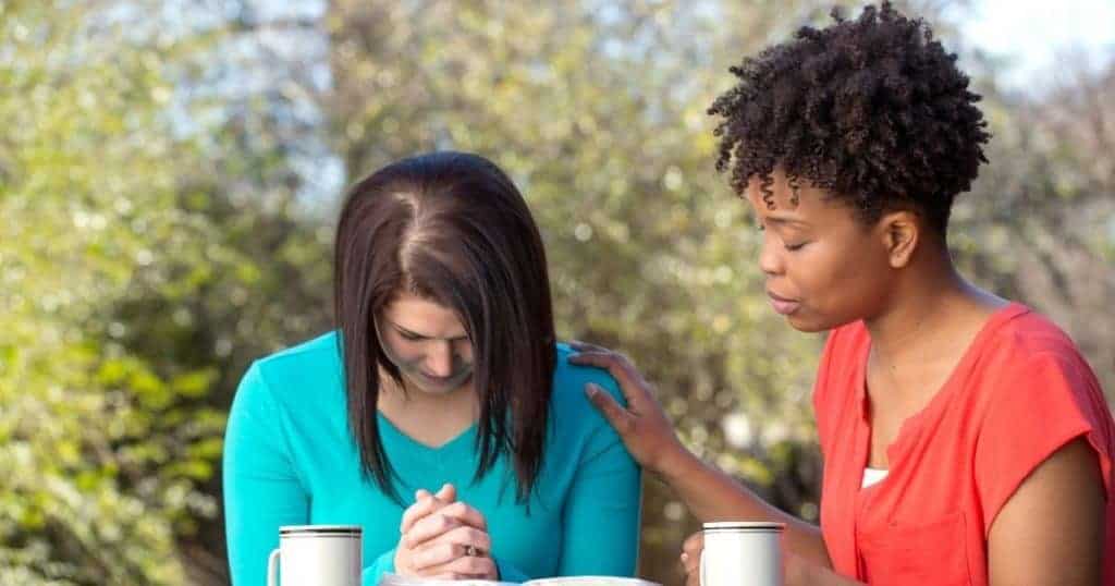 two women praying together at a table outside